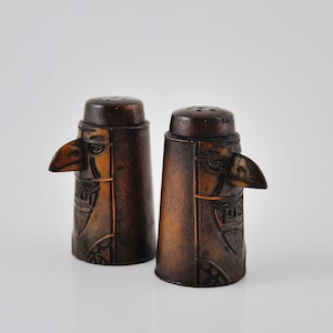 May include: A pair of brown salt and pepper shakers shaped like stylised human faces with bird beaks. The shakers have a carved design and are made of wood.