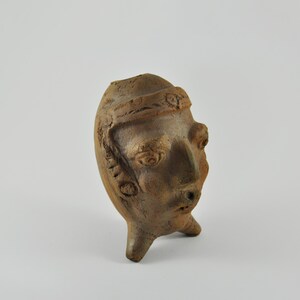 Peut inclure: Sculpture en céramique marron représentant une tête humaine avec des détails du visage et une coiffe. La sculpture a une texture rugueuse et est soutenue par trois petits pieds.
