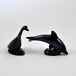 May include: Two ceramic figurines, a black goose and a black dolphin, are displayed against a white background. The goose is standing upright with its head tilted to the side, while the dolphin is leaping out of the water with its tail curved.
