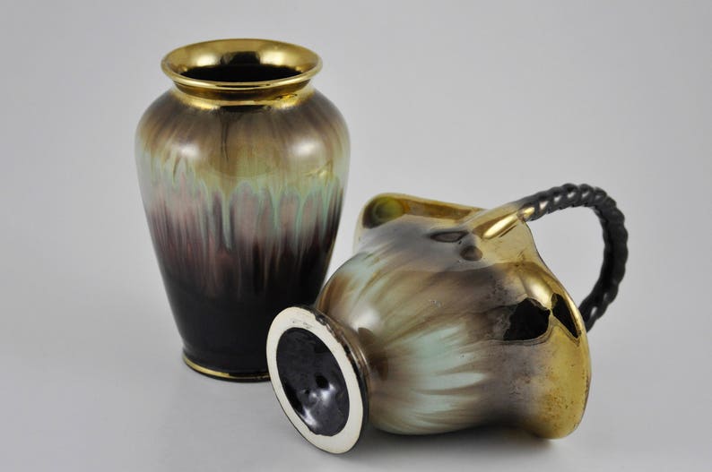 May include: Two ceramic vases with a gold, green and brown glaze. The larger vase is taller and has a rounder shape. The smaller vase has a handle and a more angular shape.