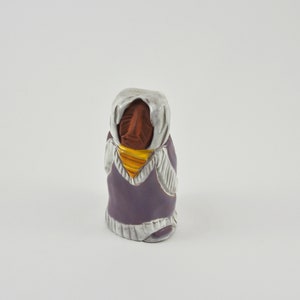 May include: A ceramic figurine of a person wearing a purple hooded garment with white trim and a yellow scarf. The figurine has a brown face and is standing on a white surface.