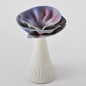 May include: A white ceramic vase with a fluted design, topped with a large, layered flower made of blue and purple fabric.