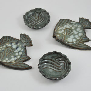 May include: Four ceramic dishes in the shape of fish and a round dish with a wave design. The dishes are glazed in a blue-green colour with white accents.