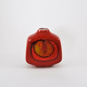 May include: A red ceramic vase with a square body and a rounded top. The vase has a yellow and orange abstract design on the front.
