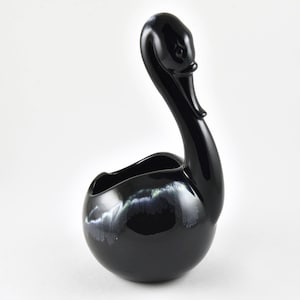 May include: Black ceramic swan planter with a white and blue glaze. The planter is shaped like a swan with its neck curved up and its head tilted to the side. The planter has a wide, open mouth that can be used to hold plants or other decorative items.
