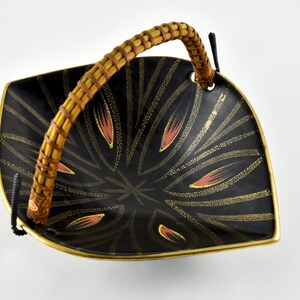 May include: A black ceramic serving dish with a woven bamboo handle. The dish has a gold and pink geometric design.