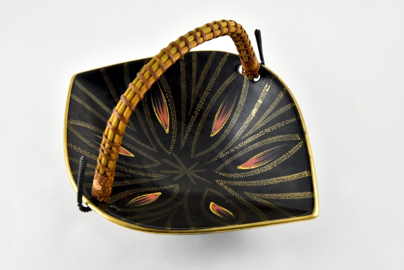 May include: A black ceramic serving dish with a woven bamboo handle. The dish has a gold and pink geometric design.