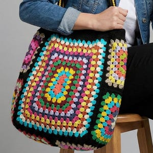Può includere: Una grande borsa tote realizzata con quadrati all'uncinetto multicolori caratterizzati da una vivace tavolozza di filati rosa, gialli, blu, verdi e viola. La borsa ha una base nera e una spessa tracolla nera con una striscia centrale gialla.