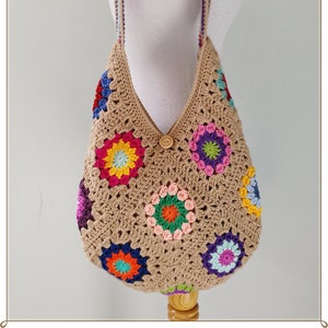 Può includere: Una borsa a tracolla all'uncinetto con una base marrone e motivi floreali colorati. La borsa ha una chiusura a bottone singola e una tracolla lunga.