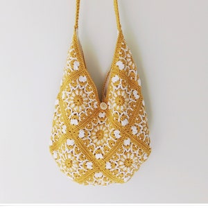 May include: A yellow crocheted tote bag with white floral designs. The bag has a long strap and a button closure.