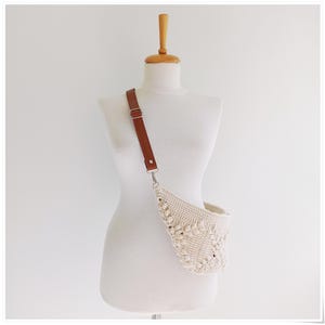May include: A white crocheted fanny pack with a brown leather strap. The bag is worn on a white mannequin.