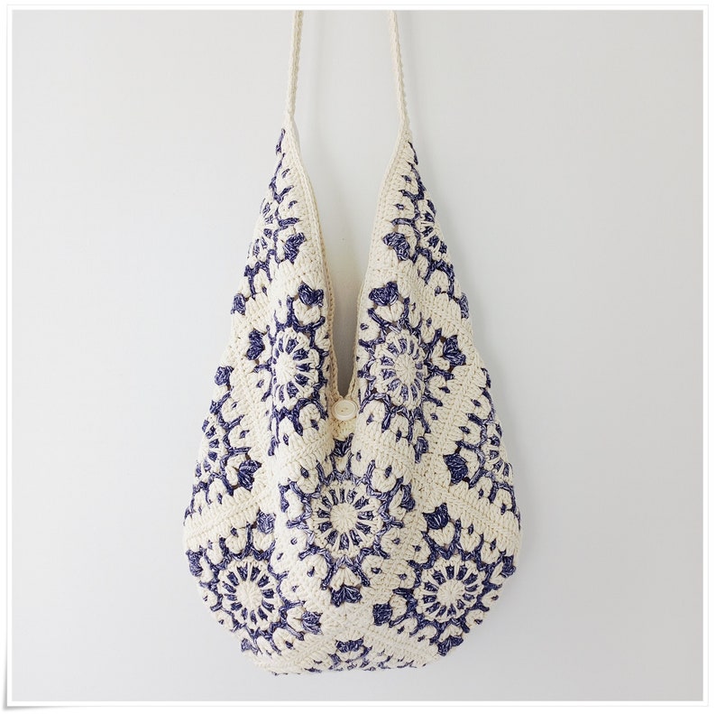 Puede incluir: Un bolso tote de crochet blanco con un patr&oacute;n floral azul. El bolso tiene un cierre de bot&oacute;n &uacute;nico y una correa larga.