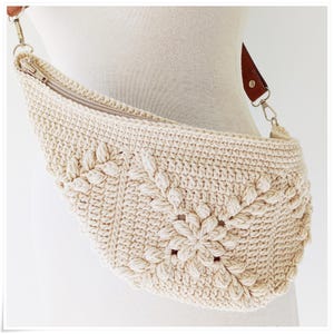May include: A crocheted cream-colored fanny pack with a brown leather strap and a silver zipper. The bag has a floral design.