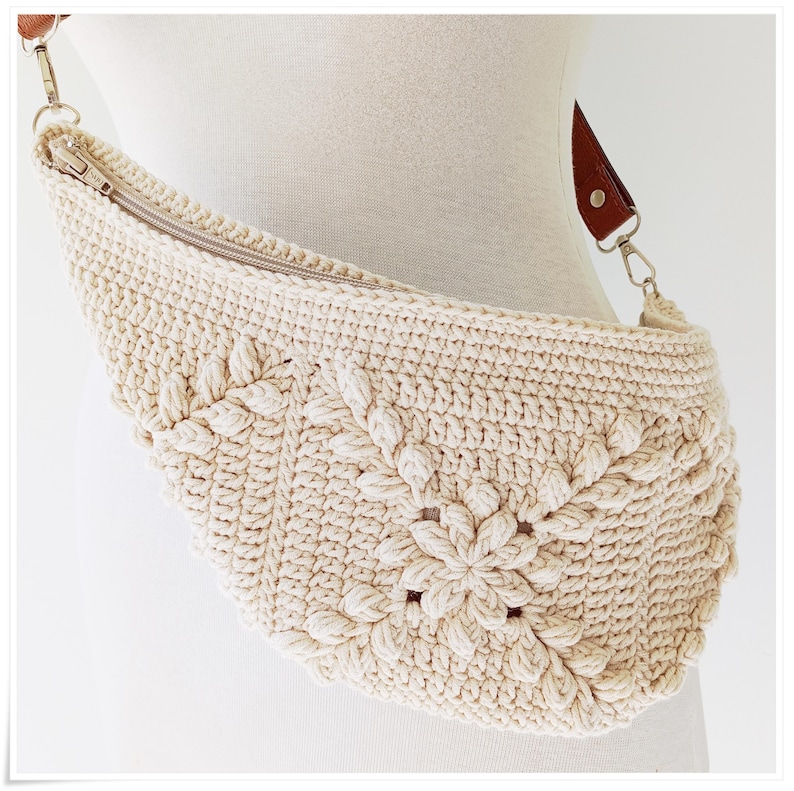 May include: A crocheted cream-colored fanny pack with a brown leather strap and a silver zipper. The bag has a floral design.