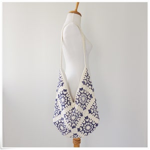 Puede incluir: Un bolso tote de crochet blanco con un patr&oacute;n floral azul. El bolso tiene una correa larga y est&aacute; colgado de un maniqu&iacute; de madera.