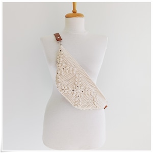 May include: A white crocheted fanny pack with a brown leather strap. The bag is worn on a white mannequin.