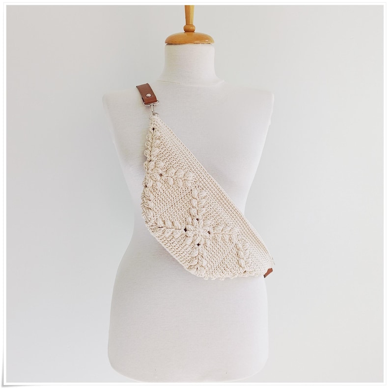 May include: A white crocheted fanny pack with a brown leather strap. The bag is worn on a white mannequin.