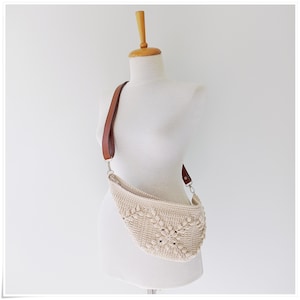 May include: A crocheted cream-colored bag with a brown leather strap. The bag is shaped like a crescent moon and has a unique woven pattern.
