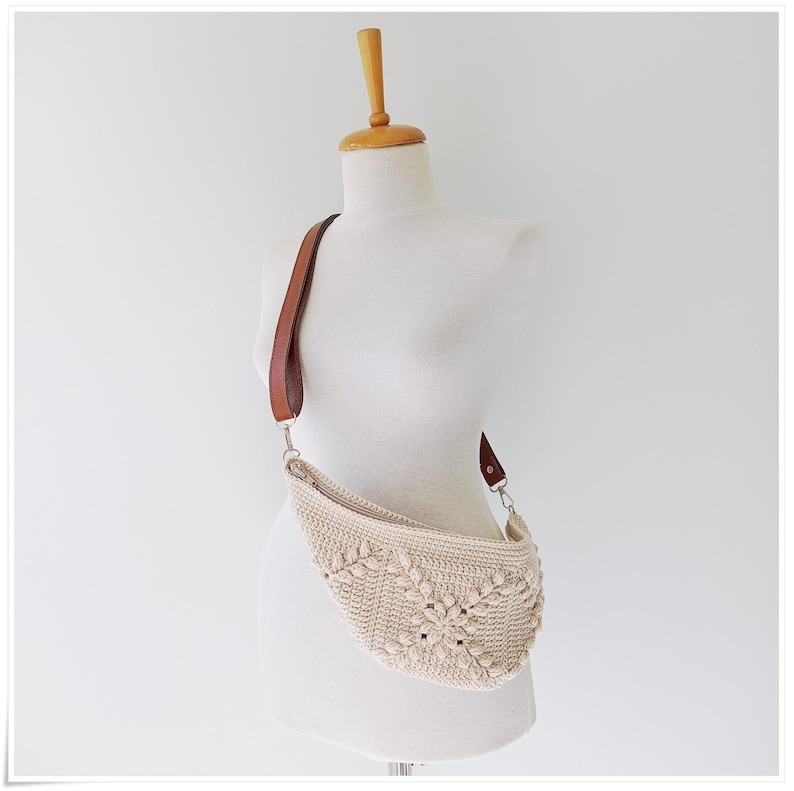 May include: A crocheted cream-colored bag with a brown leather strap. The bag is shaped like a crescent moon and has a unique woven pattern.