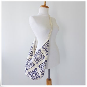 Puede incluir: Un bolso de hombro de crochet blanco con un patr&oacute;n floral azul. El bolso est&aacute; colgado de un maniqu&iacute; blanco.