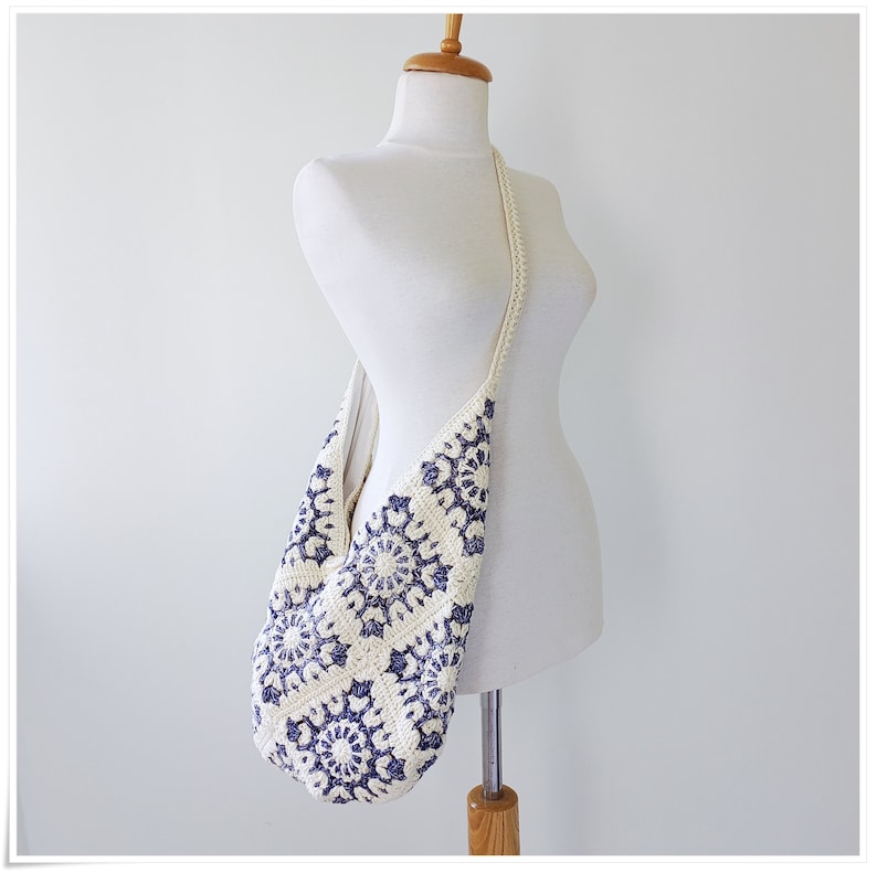 Puede incluir: Un bolso de hombro de crochet blanco con un patr&oacute;n floral azul. El bolso est&aacute; colgado de un maniqu&iacute; blanco.