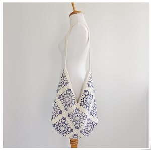 Puede incluir: Un bolso tote de crochet blanco con un patr&oacute;n floral azul. El bolso tiene una correa larga y se lleva sobre el hombro.