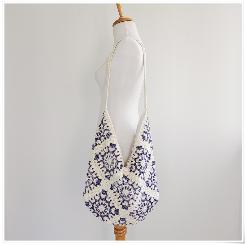 Puede incluir: Un bolso tote de crochet blanco con un patr&oacute;n floral azul. El bolso tiene una correa larga y se lleva sobre el hombro.