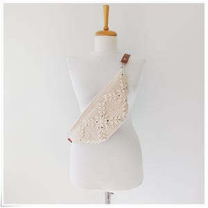 May include: A white crocheted fanny pack with a brown leather strap. The bag is worn on a white mannequin.