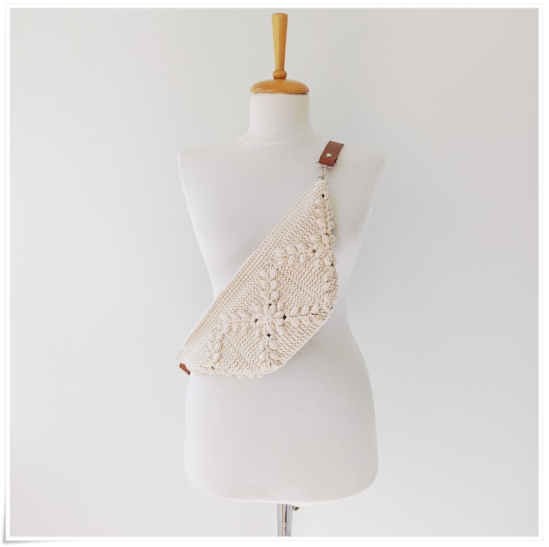 May include: A white crocheted fanny pack with a brown leather strap. The bag is worn on a white mannequin.