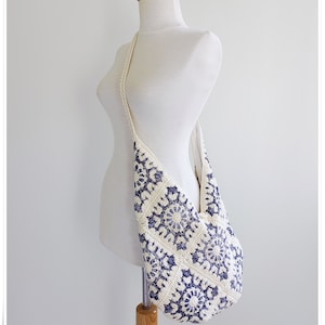 Puede incluir: Un bolso de hombro de crochet blanco con un patr&oacute;n floral azul. El bolso tiene una correa larga y est&aacute; hecho de un material suave y ligero.