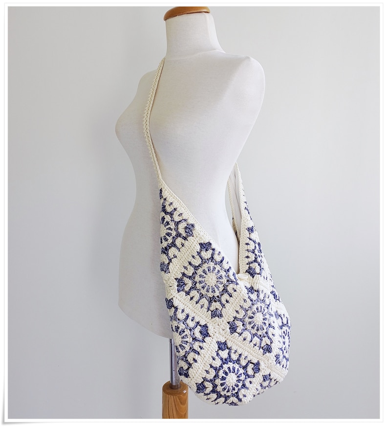 Puede incluir: Un bolso de hombro de crochet blanco con un patr&oacute;n floral azul. El bolso tiene una correa larga y est&aacute; hecho de un material suave y ligero.
