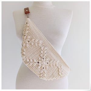 May include: A cream-colored crocheted crossbody bag with a brown leather strap. The bag has a unique floral design.