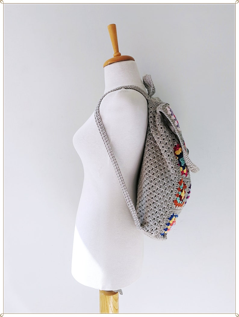 Pu&ograve; includere: Uno zaino in crochet grigio con motivi geometrici colorati sul retro. Lo zaino &egrave; su un manichino bianco.