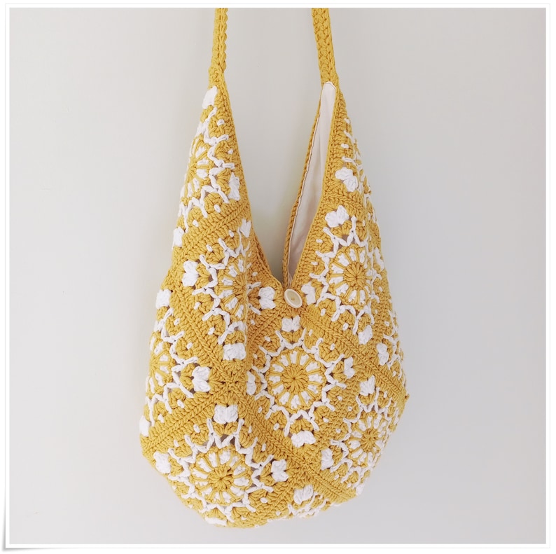 Puede incluir: Una bolsa de mano de crochet amarilla con dise&ntilde;os florales blancos. La bolsa tiene un cierre de bot&oacute;n &uacute;nico y una correa larga.