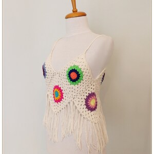 Puede incluir: Un top blanco de crochet con un dobladillo de flecos y flores de crochet de colores. El top tiene un escote en V y tirantes finos.