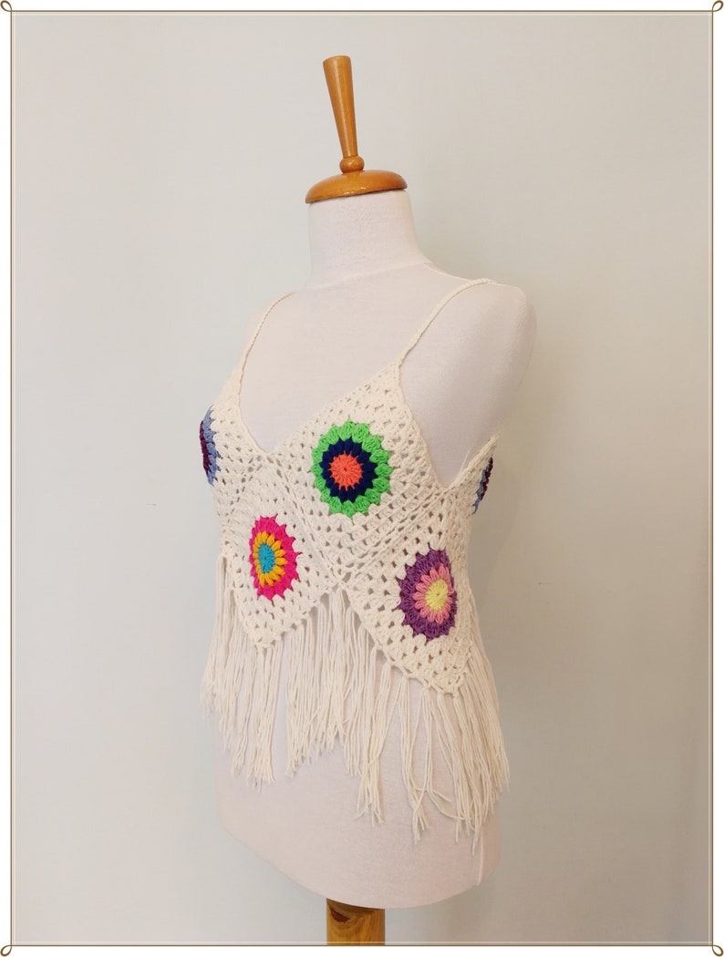 Puede incluir: Un top blanco de crochet con un dobladillo de flecos y flores de crochet de colores. El top tiene un escote en V y tirantes finos.