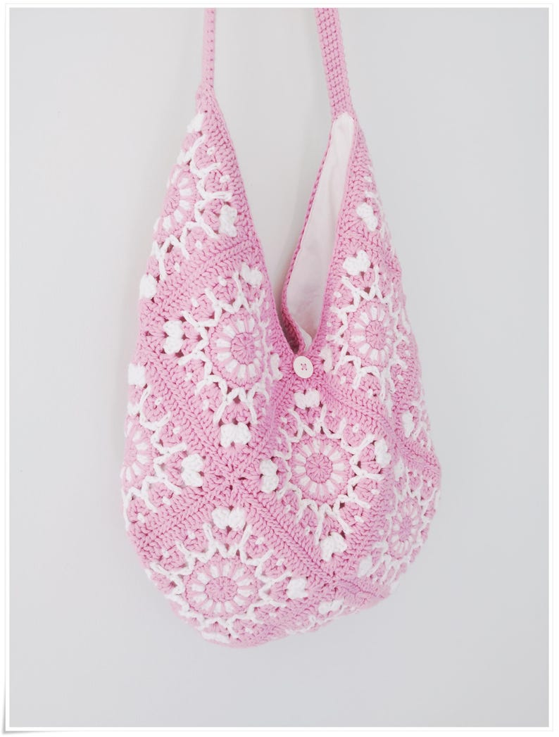 May include: A pink and white crocheted tote bag with a single button closure. The bag is made with a repeating floral pattern.