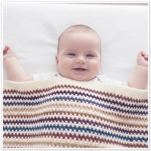 May include: A baby lying on a white surface, covered by a crocheted blanket featuring horizontal stripes in shades of cream, dark blue, and brown. The blanket has a textured, repeating stitch pattern and a solid cream-colored border along the top edge.