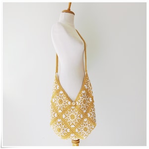 Puede incluir: Un bolso de hombro de crochet amarillo y blanco con cierre de bot&oacute;n. El bolso est&aacute; hecho de un patr&oacute;n cuadrado con un dise&ntilde;o floral.