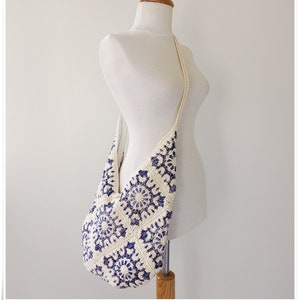 Puede incluir: Un bolso de hombro de crochet blanco con un patr&oacute;n floral azul. El bolso tiene una correa larga y se lleva en un maniqu&iacute; blanco.
