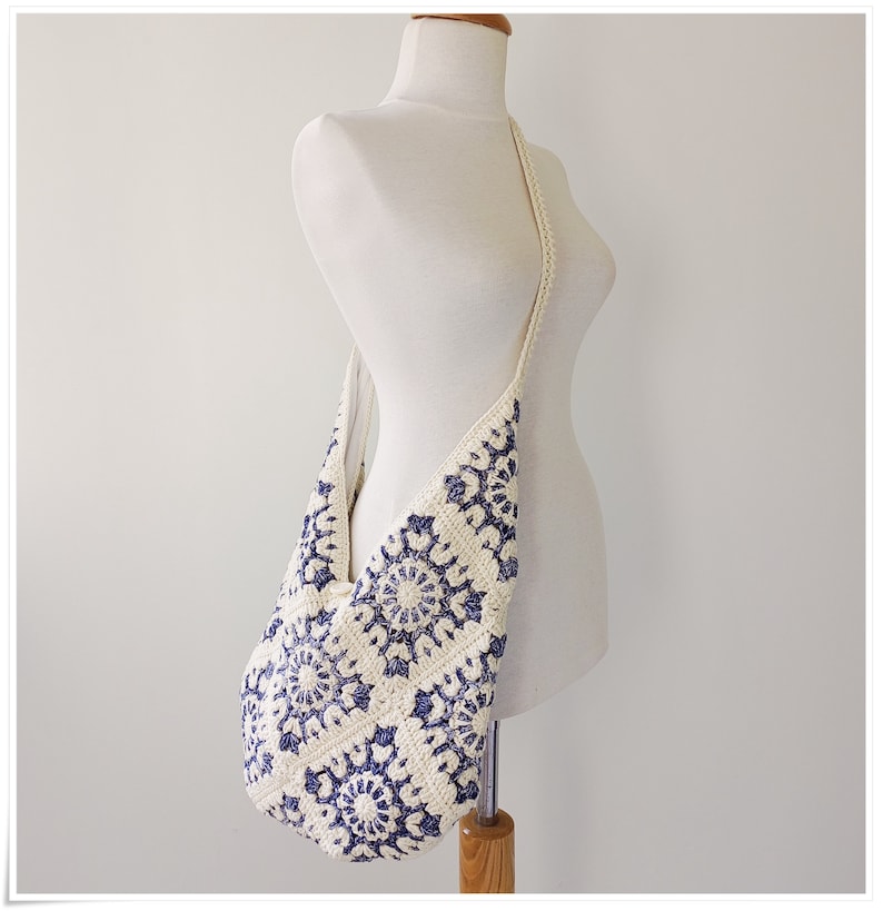 Puede incluir: Un bolso de hombro de crochet blanco con un patr&oacute;n floral azul. El bolso tiene una correa larga y se lleva en un maniqu&iacute; blanco.