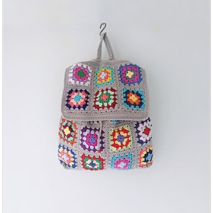 Pu&ograve; includere: Uno zaino in crochet grigio con chiusura a lembo e toppe quadrate di granny colorate. Lo zaino &egrave; appeso a un gancio contro un muro bianco.