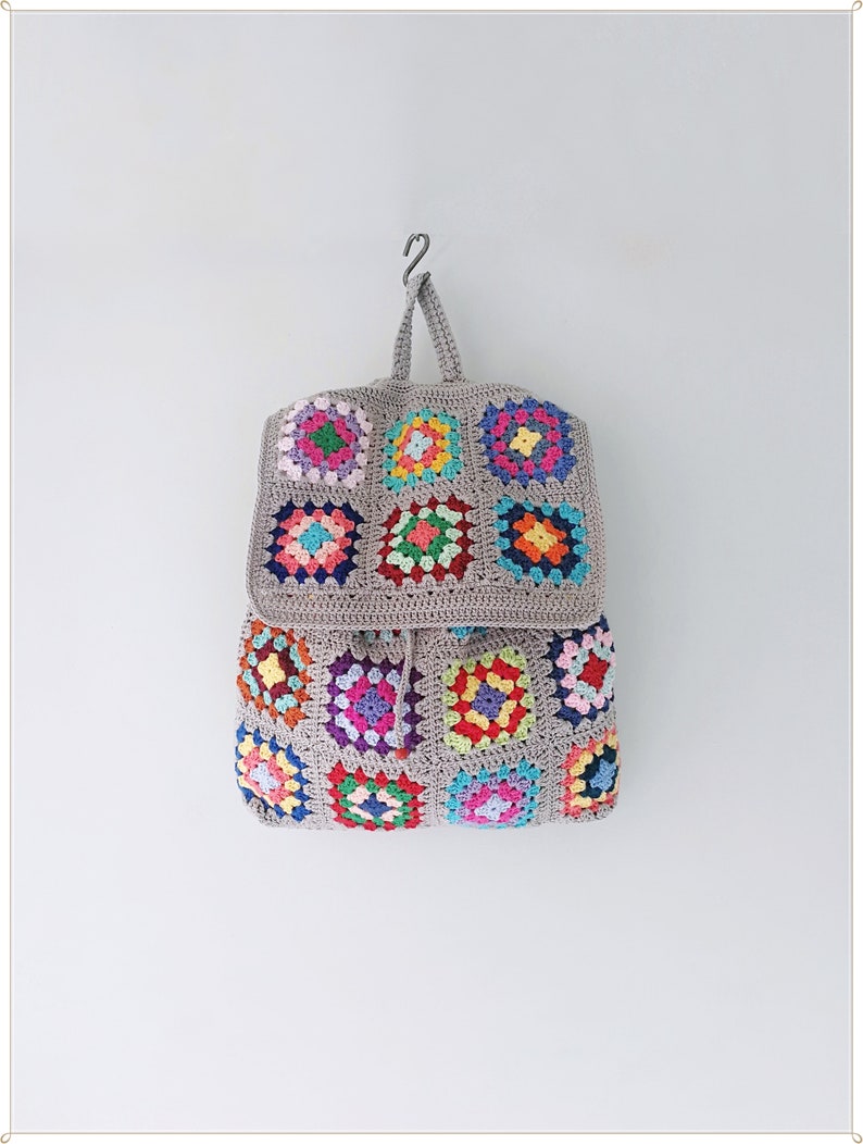 Pu&ograve; includere: Uno zaino in crochet grigio con chiusura a lembo e toppe quadrate di granny colorate. Lo zaino &egrave; appeso a un gancio contro un muro bianco.