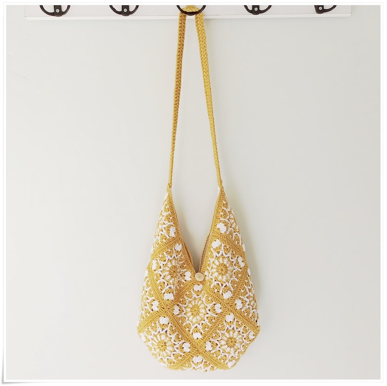 Puede incluir: Un bolso de hombro de crochet amarillo con detalles florales blancos. El bolso tiene una correa larga y un cierre de bot&oacute;n.