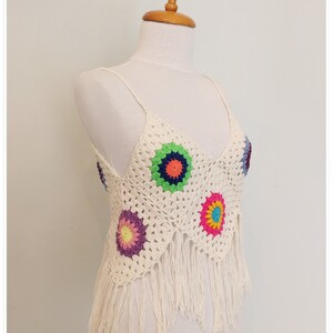 Puede incluir: Top blanco de crochet con detalles de flores de colores y ribete de flecos.