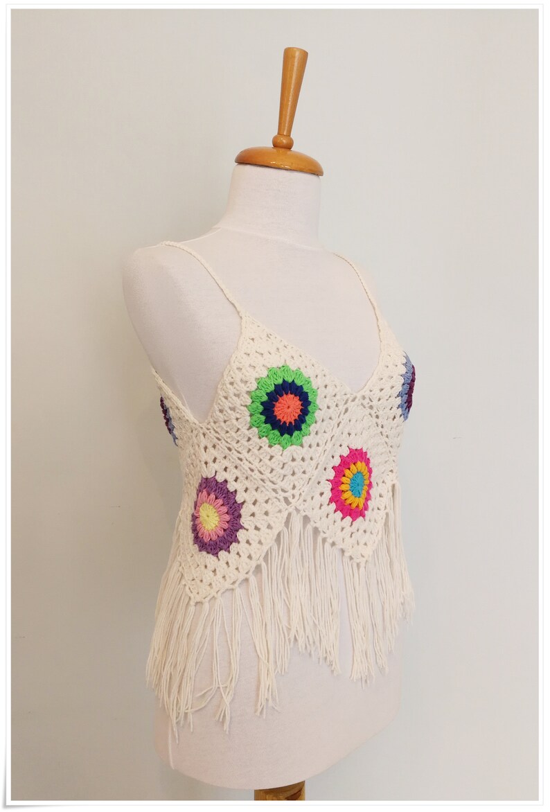 Puede incluir: Top blanco de crochet con detalles de flores de colores y ribete de flecos.