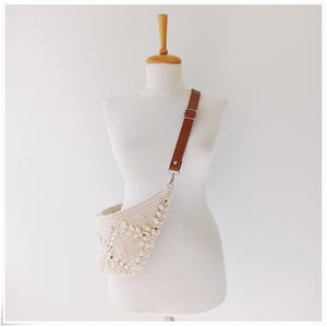 May include: A white crocheted crossbody bag with a brown leather strap. The bag has a unique, textured design.