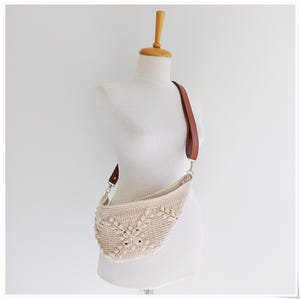 May include: A crocheted cream-colored bag with a brown leather strap. The bag is shaped like a crescent moon and has a woven pattern.