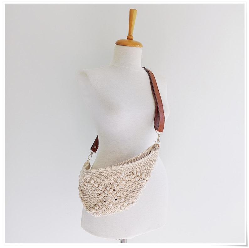 May include: A crocheted cream-colored bag with a brown leather strap. The bag is shaped like a crescent moon and has a woven pattern.