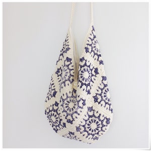 Puede incluir: Una bolsa de mano de crochet blanca con un patr&oacute;n floral azul. La bolsa tiene una correa larga y est&aacute; hecha de un hilo suave y texturizado.